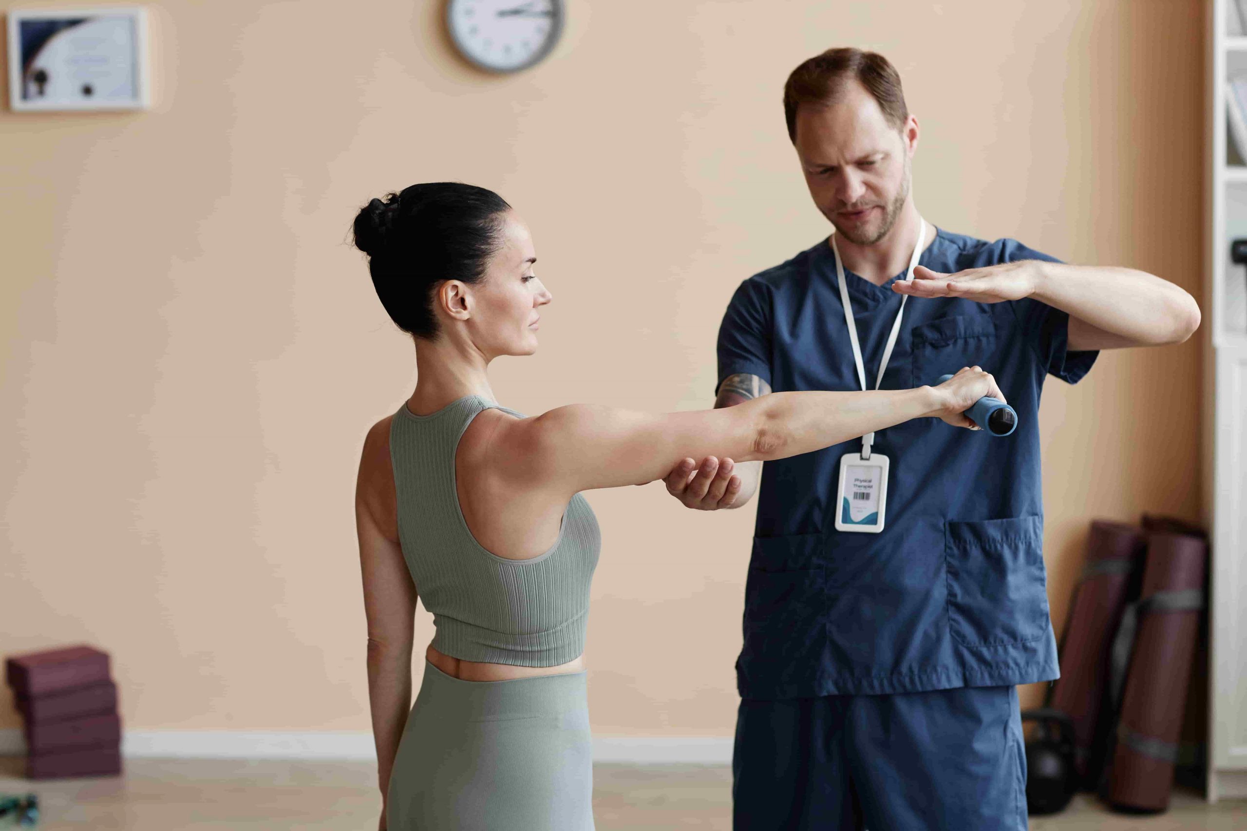 Fisioterapeuta de Clínica Trajano guía a una paciente mientras realiza un ejercicio de movilización de hombro con una barra ligera en la sala de tratamiento.